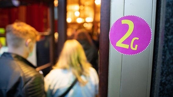 Ein Aufkleber mit der Aufschrift "2G" ist am Eingang eines Gebäudes zu sehen, zwei Menschen betreten es durch eine Tür. © dpa bildfunk Foto: Daniel Reinhardt