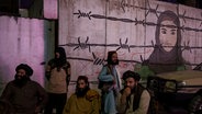 Taliban sitzen vor einer Mauer, auf der eine Frau hinter Stacheldraht abgebildet ist. © picture alliance / ASSOCIATED PRESS | Felipe Dana 