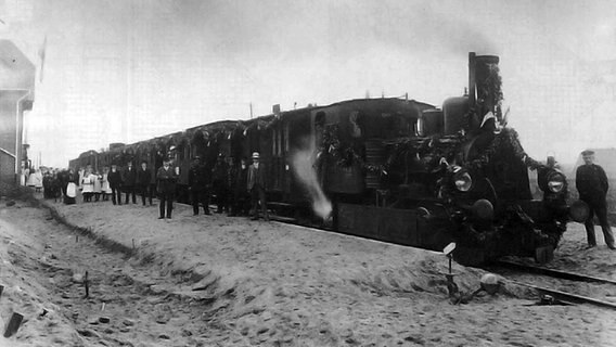 Ein alter Zug ist mit Girlanden geschmückt und steht im Bahnhof Bönningstedt. Daneben stehen einige Menschen. Das Foto ist schwarz-weiß und wurde ca. Anfang des 20. Jahrhunderts aufgenommen. © AKN Eisenbahn AG 