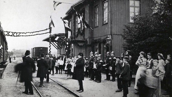 Am Quickborner Bahnhof stehen viele Menschen. Das Bahnhofsgebäude ist mit Girlanden geschmückt. Das Foto ist schwarz-weiß und wurde ca. Anfang des 20. Jahrhunderts aufgenommen. © AKN Eisenbahn AG 