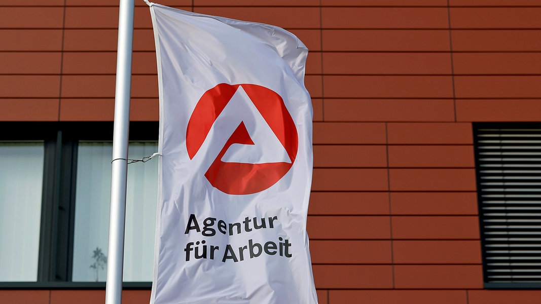 Das Logo und der Schriftzug der Agentur für Arbeit auf einer Fahne vor einem Gebäude.