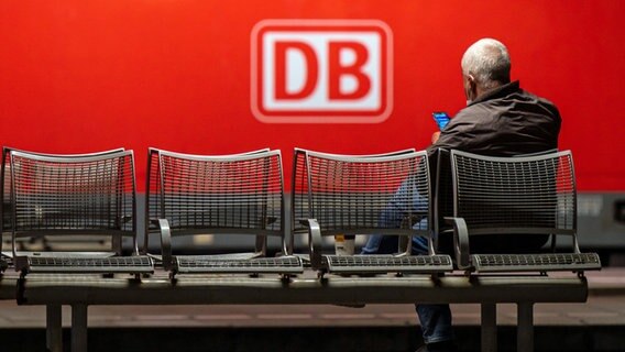Das Logo der Deutschen Bahn (DB) prangt an der Seite einer Lok, während ein Mann auf einer Bank auf einem Bahnsteig wartet. © dpa-Bildfunk 