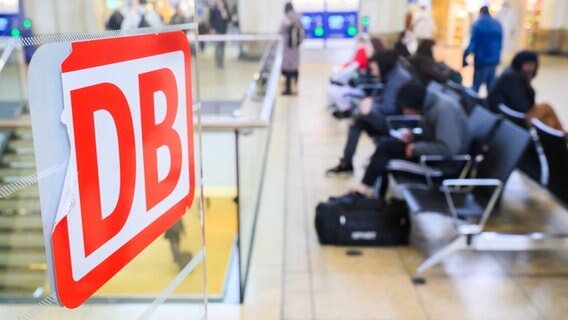 DB-Logo klebt während des Bahnstreiks an einem Bahnhof -  im Hintergrund warten Bahnkunden © Julian Stratenschulte/dpa Foto: Julian Stratenschulte/dpa