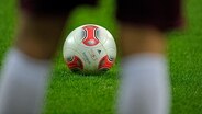 Ein Fußball liegt auf dem Rasen, fotografiert durch die Beine eines Fußballers. © dpa picture alliance Foto: MiS