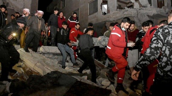 Ein von der amtlichen syrischen Nachrichtenagentur SANA veröffentlichtes Foto zeigt Rettungskräfte und Menschen, die Verletzte aus Trümmern eines zerstörten Gebäudes tragen. © SANA/dpa Foto: SANA/dpa