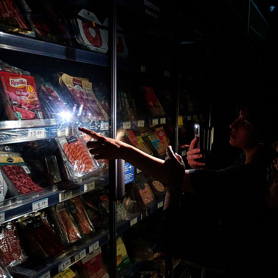 Menschen in Spanien kaufen während eines massiven Stromausfalls in einer Kühltheke im Supermarkt ein, beleuchtet durch ihre Smartphone-Kameras. © picture alliance/dpa/AP Foto: Miguel Oses