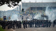 Anhänger des ehemaligen brasilianischen Präsidenten Bolsonaro (hinten) geraten in der Hauptstadt Brasilia mit Ordnungskräften aneinander, die hinter Absperrungen eine Kette bilden und Tränengasgranaten auf die Demonstranten abfeuern. © dpa bildfunk Foto: Matheus Alves