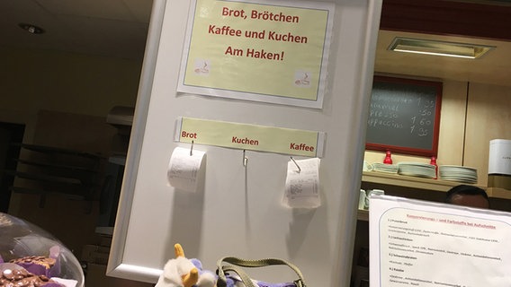 Auf einem Schild steht "Brot, Brötchen, Kaffee und Kuchen am Haken!" - Hier können Kunden im Voraus bezahlte Snacks verschenken. © NDR Foto: Lennart Seebald