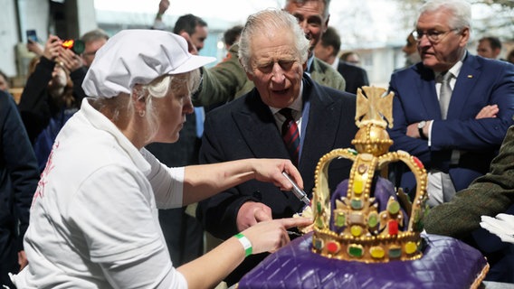 König Charles III. von Großbritannien (M) schneidet im Ökodorf Brodowin eine Torte in Form einer Krone an, die speziell für seinen Besuch angefertigt wurde. © dpa-Pool/dpa Foto: Jens Büttner