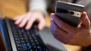 Ein Mann sitzt am Laptop und hält ein Smartphone mit in der Hand. © Monika Skolimowska/dpa 