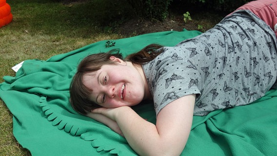 Eine junge erwachsene Frau, die das Downsyndrom hat, liegt auf einer Decke auf einer Wiese. © KIDS Hamburg 