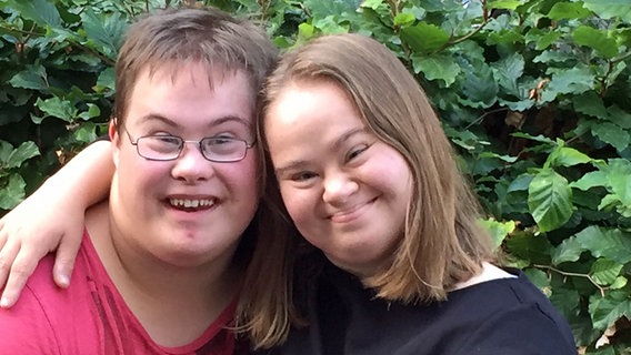 Zwei junge Erwachsene, die das Downsyndrom haben, halten sich Arm in Arm fest und lächeln in die Kamera. © KIDS Hamburg 
