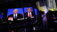 Bilder von US-Präsident Joe Biden und seinem republikanischen Konkurrenten Donald Trump spiegeln sich in einer offenen Autotür, während Zuschauer eine TV-Wahlkampfdebatte auf einer riesigen Leinwand in einem Freiluftkino verfolgen. © Rebecca Blackwell/AP Foto: Rebecca Blackwell/AP