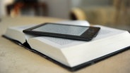 Ein E-Book-Reader liegt auf einem aufgeschlagenen Buch. © dpa picture alliance Foto: Jens Kalaene