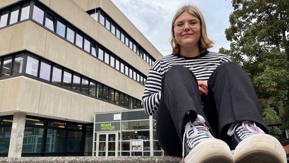 Eva, Studentin in Hamburg, sitzt auf dem Campus auf einer Mauer. Im Hintergrund ist ein Universitätsgebäude und die Mensa "Blattwerk" zu sehen. Eva, Studentin in Hamburg, sitzt auf dem Campus auf einer Mauer. Im Hintergrund ist ein Universitätsgebäude und die Mensa "Blattwerk" zu sehen. © NDR Info Foto: Lisa Hentschel
