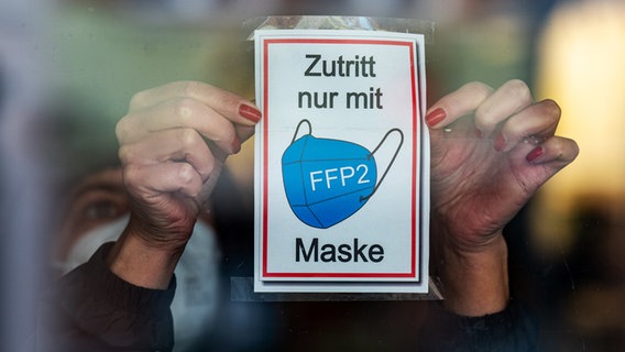 Eine Frau klebt ein Hinweisschild mit der Aufschrift "Zutritt nur mit FFP2-Maske" an eine Glastür am Eingang eines Geschäfts. © picture alliance/dpa | Armin Weigel 