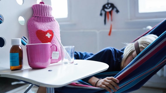 Fieberthermometer, Hustensaft, Wärmflasche und eine Tasse Tee stehen auf einem Stuhl, während ein Kind im Hintergrund in einer Hängematte liegt. © picture alliance/dpa | Annette Riedl Foto: Annette Riedl