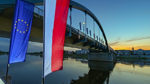 Die Fahne der Europäischen Union und die Nationalflagge Polens wehen im Wind zum Sonnenaufgang vor der Stadtbrücke über den Grenzfluss Oder zwischen Frankfurt (Oder) und dem polnischen Slubice. © Patrick Pleul/dpa 