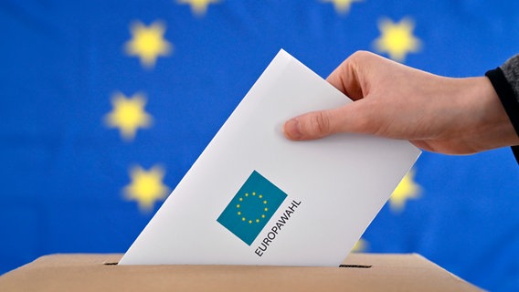 Ein Wahlbrief wird in eine Wahlurne gesteckt, im Hintergrund eine EU-Flagge. © picture alliance / Panama Pictures 
