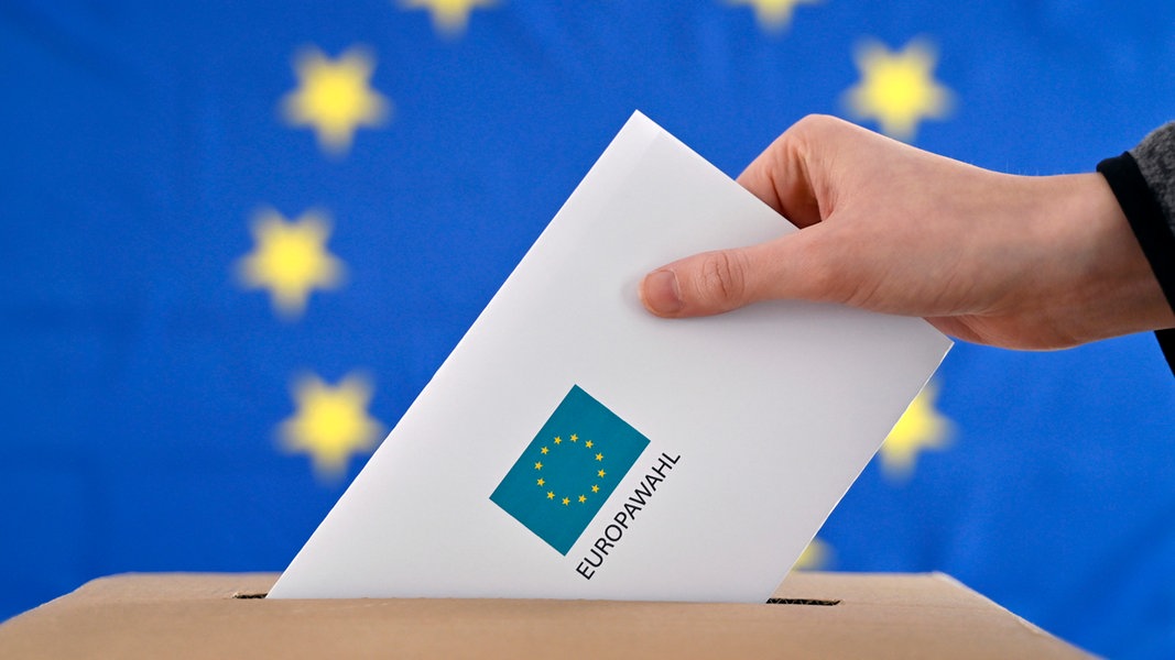 Ein Wahlbrief wird in eine Wahlurne gesteckt, im Hintergrund eine EU-Flagge.