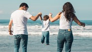 Ein Elternpaar hält sein kleines Kind am Strand an den Händen und lässt es fliegen © Colorbox Foto: Oleksandr Latkun