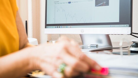 Die Hand einer Frau ist an einem Schreibtisch zu sehen, auf dem ein PC steht, der Aktien-Charts anzeigt. © picture alliance / dpa-tmn Foto: Christin Klose