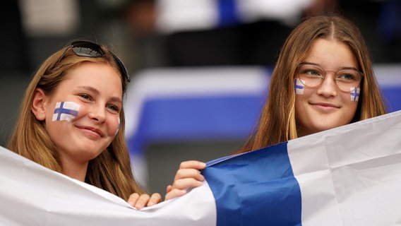 Zwei Mädchen halten lächelnd eine finnische Flagge hoch. Auf ihren Wangen sind ebenfalls finnische Flaggen gemalt. © PA Wire/dpa Foto: Tim Goode