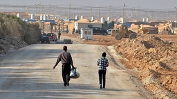 Syrische Flüchtlinge gehen auf einer Straße. Im Hintergrund ein Auffanglager in Jordanien. Syrische Flüchtlinge gehen auf einer Straße. Im Hintergrund ein Auffanglager in Jordanien. © dpa picture alliance Foto: Yann Foreix