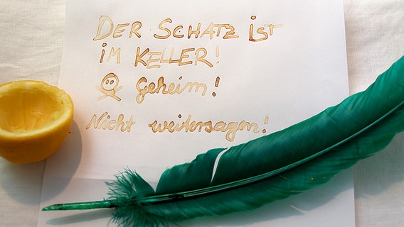 Eine Feder und eine Zitrone mit einem Blatt Papier, auf dem mit Geheimtinte geschrieben steht "Der Schatz ist im Keller! Geheim! © NDR Foto: Katharina Mahrenholtz