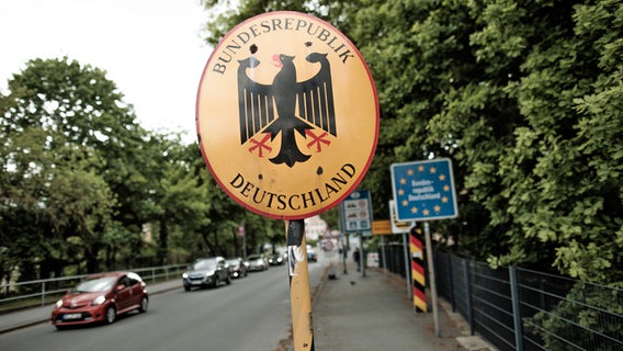 Ein Schild mit der Aufschrift Bundesrepublik Deutschland und dem Bundesadler, Landesschild der Bundesrepublik Deutschland mit den Sternen der Europäischen Union auf blauem Grund, ein deutscher Grenzpfosten und das Ortseingangsschild vor der Zufahrt zur Einreisekontrolle am deutsch-polnischen Grenzübergang Görlitz. © picture alliance/dpa | Carsten Koall 