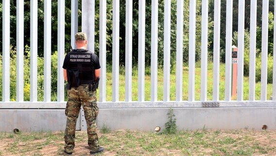 Ein polnischer Grenzsoldat steht vor einem Grenzzaun an der Grenze zu Belarus, dahinter ist eine Belarussische Grenzmarkierung zu sehen. © IMAGO / Eastnews 