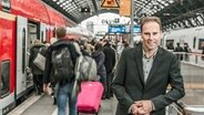 Hagen Lesch steht auf einem Bahnsteig, Reisende steigen in einen Zug ein und aus. © Institut der Deutschen Wirtschaft, Köln 