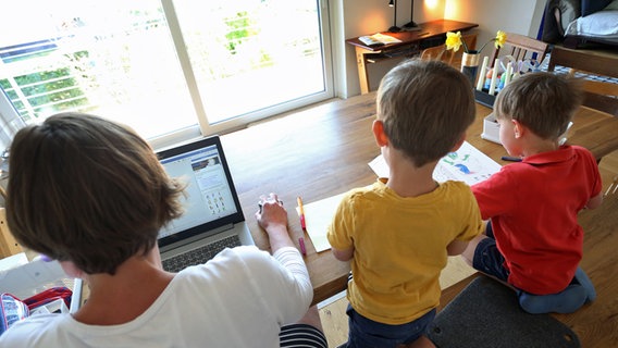 Eine arbeitet zu Hause an einem Laptop, während ihre Kinder neben ihr malen und ein Buch ansehen. © picture alliance/dpa Foto: Karl-Josef Hildenbrand