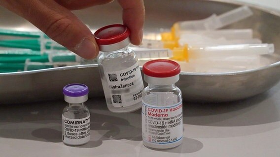 Eine Hand nimmt eine Impfdose vom Impfstoff AstraZeneca vom Tisch. Links im Bild steht eine Impfdose vom Impfstoff Biontech, rechts im Bild steht eine Impfdose vom Impfstoff Moderna. © dpa bildfunk Foto: Till Simon Nagel