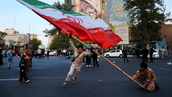 15.04.2024, Iran, Teheran: Demonstranten schwenken eine riesige iranische Flagge bei einer anti-israelischen Versammlung vor einem anti-israelischen Banner an der Wand eines Gebäudes auf dem Felestin (Palästina) Platz. © dpa Foto: Vahid Salemi