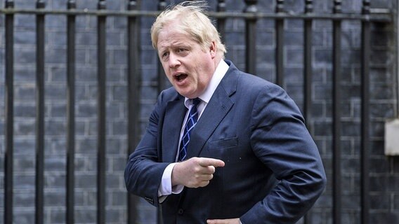 Der britische Politiker Boris Johnson geht an einem Gebäude mit dunkler Fassade vorbei und zeigt mit einem Finger zur Seite. © dpa picture alliance/Photoshot 