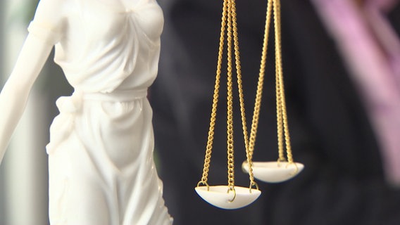 Eine weiße Justitia-Plastik (Göttin der Gerechtigkeit) mit zwei Waagschalen an goldenen Kettchen in Nahaufnahme © ndr.de 
