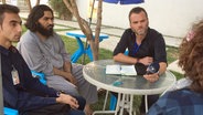 ARD-Korrespondent Jürgen Webermann (M.) sitzt beim Interview mit zwei afghanischen Studenten und einem Dolmetscher an einem Tisch. © NDR 