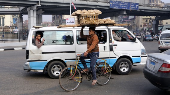 A young man on a bicycle balances a wooden tray full of freshly baked bread on his head in the busy traffic of Cairo.  © dpa picture alliance Photo: Matthias Toedt