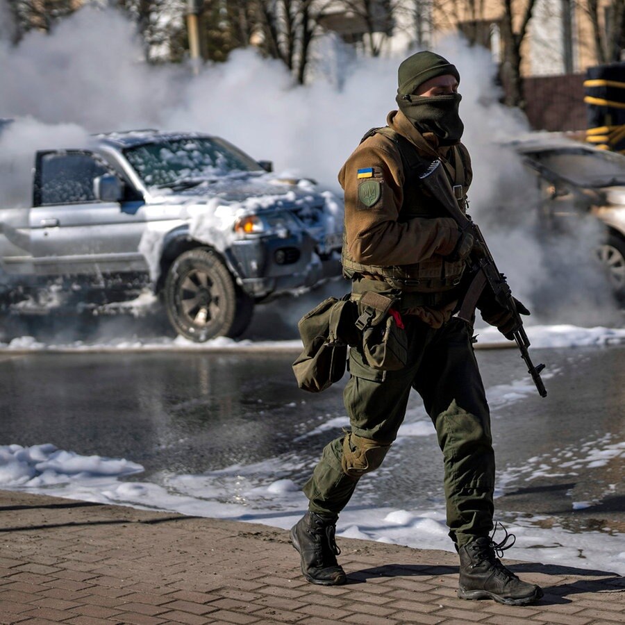 Ein ukrainischer Soldat bezieht Stellung vor einer Militäreinrichtung in Kiew, während Feuerwehrleute im Hintergrund zwei brennende Autos löschen. © dpa-Bildfunk 