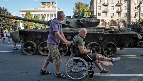 Ukraine, Kiew: Ein Mann schiebt einen verwundeten ukrainischen Soldaten in einem Rollstuhl an einem zerstörten russischen Panzer vorbei. © AP/dpa Foto: Evgeniy Maloletka