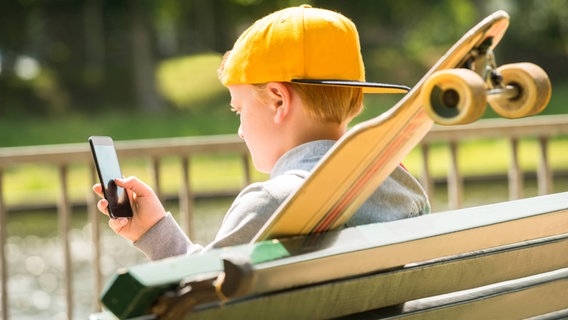 Ein Kind mit einer gelben Baseball-Cap sitzt auf einer Parkbank und schaut auf ein Smartphone © picture alliance / PantherMedia Foto: Andriy Popov