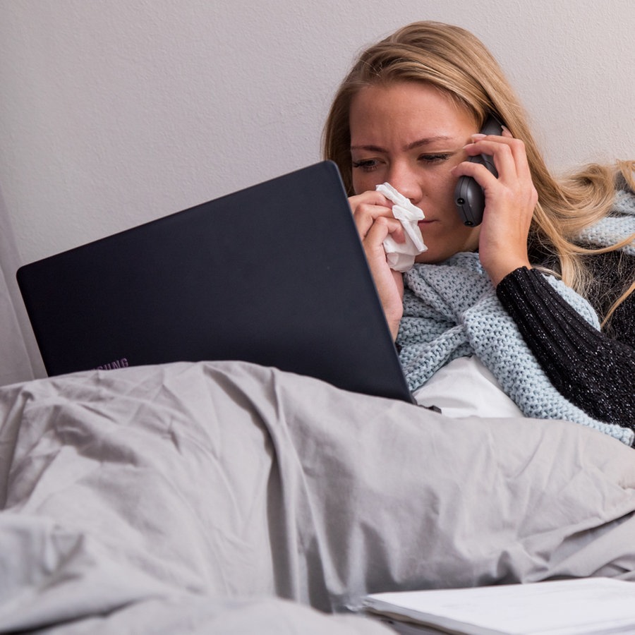 Eine Frau liegt krank im Bett. Telefoniert mit einem Laptop auf den Knien und putzt sich die Nase © picture alliance / Christin Klose Foto: Christin Klose
