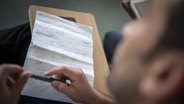 Ein Migrant hält einen Kugelschreiber in der Hand, vor ihm liegt ein Zettel voller Notizen. © imago/photothek Foto: Florian Gaertner