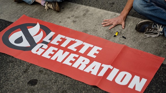Aktivisten der "Letzten Generation" haben sich auf einer Straße festgeklebt, vor ihnen liegt ein Transparent mit dem Logo der Gruppe. © dpa-Bildfunk Foto: Swen Pförtner