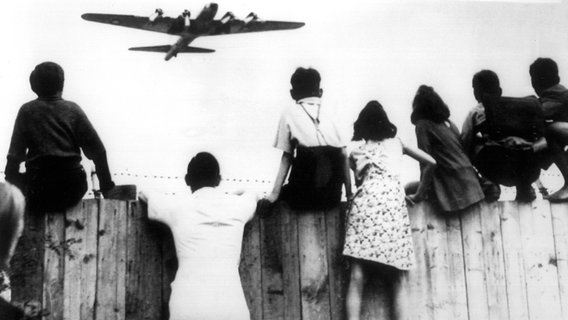Im Juni 1948 fliegt ein Flugzeug im Landeanflug in Berlin tief über ein paar Menschen hinweg, die nach oben schauen. (Historisches Schwarzweiß-Bild) © imago 
