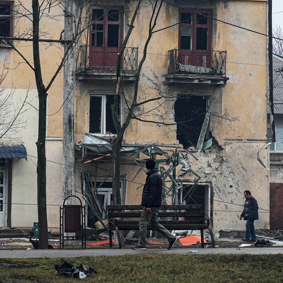 Menschen gehen auf einer Straße der ukrainischen Stadt Mariupol an einem Wohnhaus vorbei, das durch Beschuss beschädigt ist. © Evgeniy Maloletka/AP/dpa 
