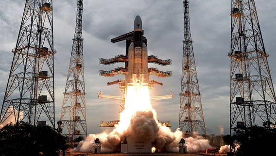 Eine indische Trägerrakete startet ihren Flug ins All Richtung Mond. Um die Rakete herum stehen Masten. Durch den Antrieb der Rakete entstehen Rauchwolken. © dpa picture alliance Foto: //