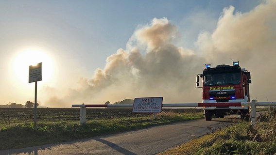 Ein Löschfahrzeug der Feuerwehr steht an einer Schranke zu einem Bundeswehr-Gelände. Im Hintergrund ist Rauch im Sonnenlicht über einem Stück Moorlandschaft zu sehen. © dpa-Bildfunk Foto: Stephan Konjer