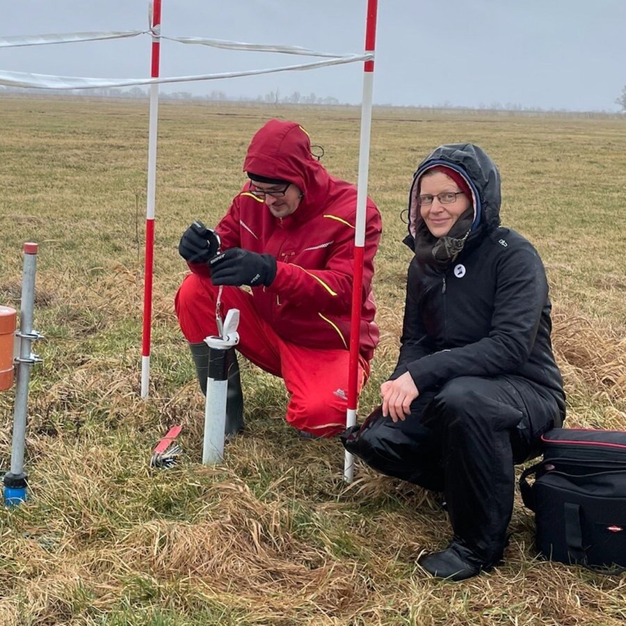 Eine Frau und ein Mann hocken auf einem Feld vor Gerätschaften. © NDR Info Foto: Nicole Ahles